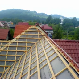 W trakcie budowy więźby dachowej z widocznymi łatami, kontrłatami i membraną, z fragmentem ułożonej dachówki ceramicznej w tle oraz pracownikiem na dachu i krajobrazem górskim w oddali.
