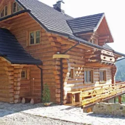 Drewniany dom z bali z ciemną dachówką, balkonem i kamiennym podmurówką na tle gór.