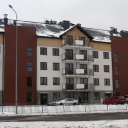 Nowoczesny, wielopiętrowy budynek z białymi ścianami i brązowymi akcentami elewacji. Balkony z czarnymi barierkami. Częściowo ośnieżony dach i otoczenie.