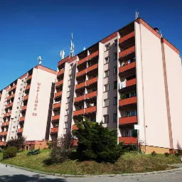 Wielopiętrowy blok mieszkalny z balkonami, ocieplony i pomalowany na jasny kolor, z widocznym napisem 'WAPIENNA 30' na elewacji, na tle błękitnego nieba.