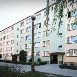 Długi blok mieszkalny z kremowo-zieloną elewacją i rzędami okien, oświetlony latarnią. Widoczny fragment chodnika i drzewa.