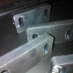 Metalowe, prostokątne elementy z owalnymi otworami, leżące na ciemnej, prawdopodobnie metalowej powierzchni warsztatowej. Na jednym z elementów widoczna linia narysowana markerem.