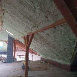 Paweł Piekielnik - Poddasze domu w trakcie prac izolacyjnych, pokryte pianką poliuretanową, widoczne drewniane elementy konstrukcyjne dachu i fragment komina z cegły. Na podłodze resztki materiałów budowlanych...