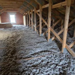 Poddasze z drewnianą konstrukcją więźby dachowej, pokryte warstwą izolacji z włókien mineralnych, widoczne okno w murze z cegły.