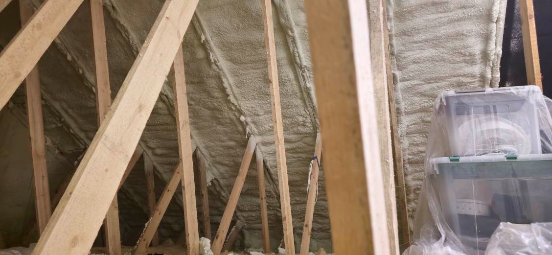 Poddasze z drewnianą konstrukcją krokwi pokryte warstwą jasnej pianki izolacyjnej, w tle widoczne przezroczyste plastikowe pojemniki.