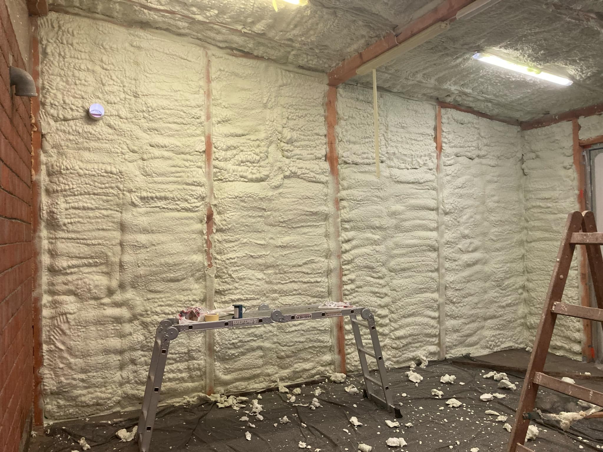 Wnętrze budynku w trakcie ocieplania pianką poliuretanową, widoczne fragmenty muru z cegły, aluminiowa drabina rozstawna i drewniana drabina przystawna, podłoga zabezpieczona folią z resztkami pianki.