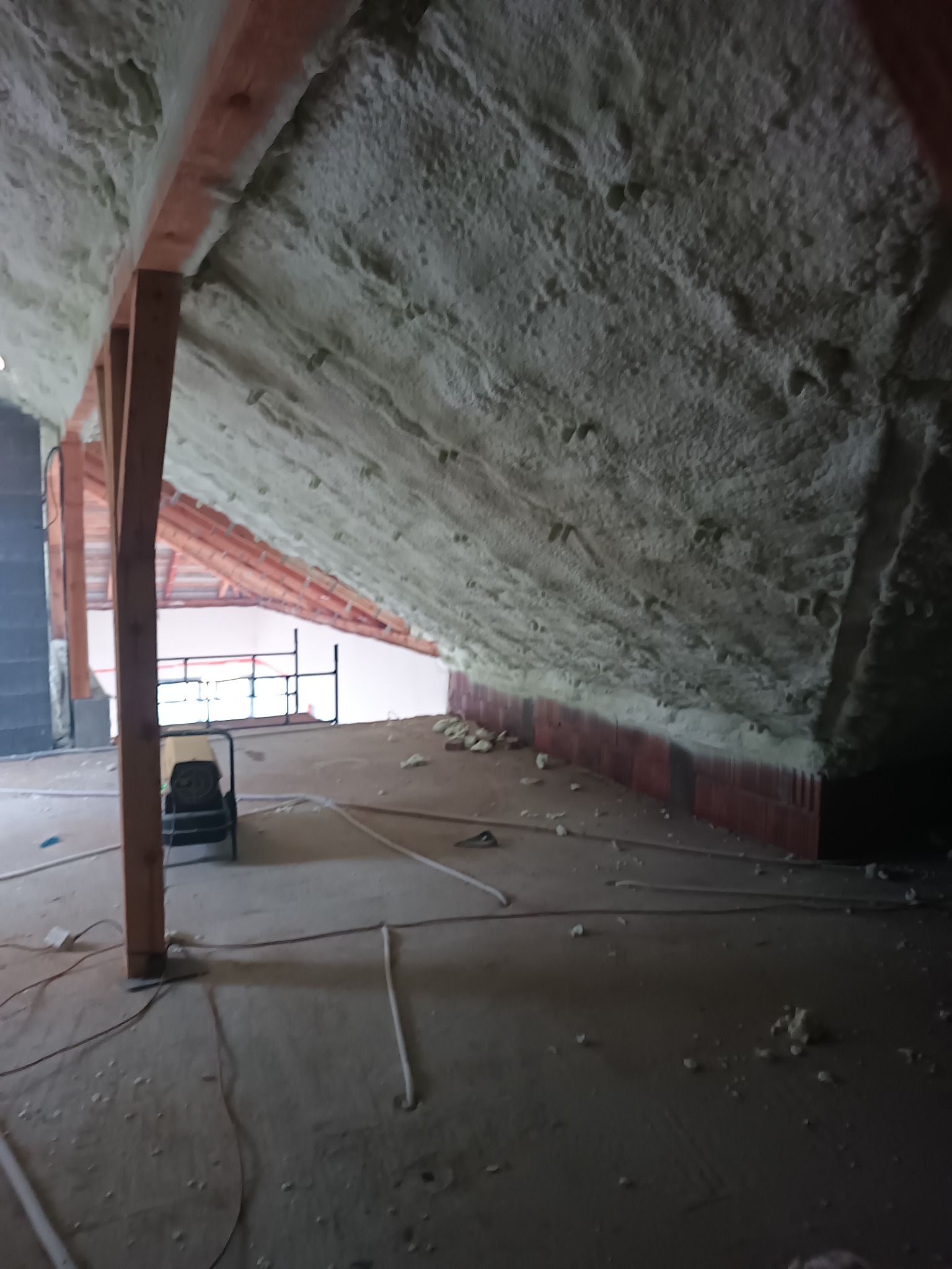 Poddasze w trakcie ocieplania pianką PUR, widoczne drewniane belki konstrukcyjne, ściany pokryte pianką, na podłodze resztki materiału i przewody, w tle rusztowanie.