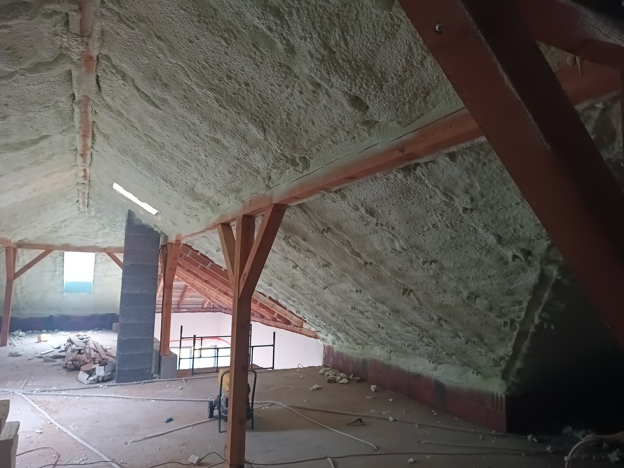 Poddasze domu w trakcie prac izolacyjnych, pokryte pianką poliuretanową, widoczne drewniane elementy konstrukcyjne dachu i fragment komina z cegły. Na podłodze resztki materiałów budowlanych...