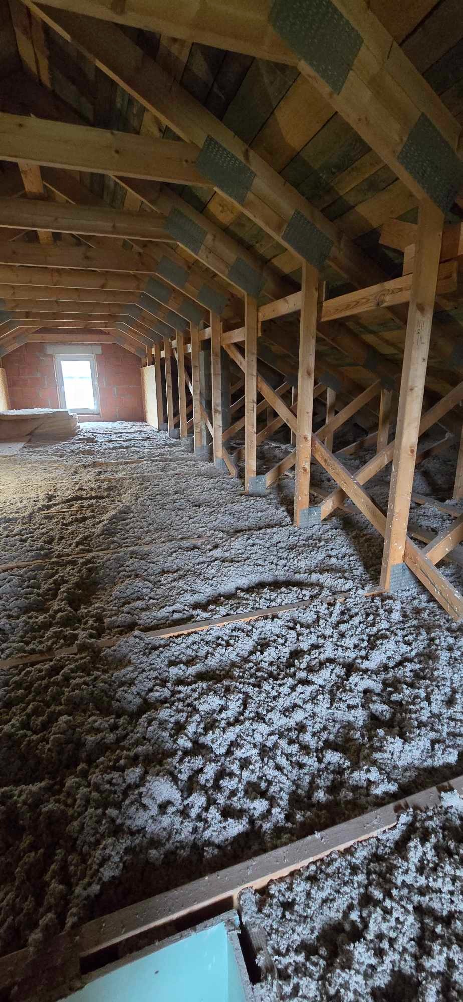 Poddasze z drewnianą konstrukcją więźby dachowej, pokryte warstwą izolacji z włókien mineralnych, widoczne okno w murze z cegły.