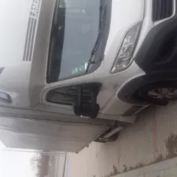 Biały bus z plandeką zaparkowany na chodniku z kostki, widok z boku na przód pojazdu, z naklejkami na przedniej szybie.