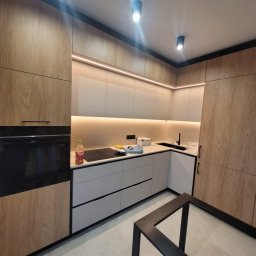 Pracownia na nowo - Nowoczesna kuchnia z drewnianymi frontami, szarymi szufladami i oświetleniem LED pod szafkami wiszącymi. Na blacie stoi butelka napoju i gąbka. Widoczny fragment metalowej konstrukcji stołu.