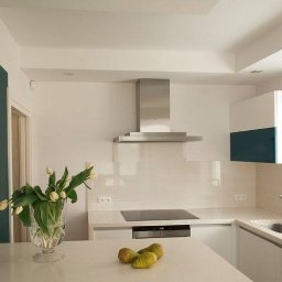 Pracownia na nowo - Nowoczesna kuchnia z białymi szafkami, blatem z kwarcytu i akcentem w postaci ściany w kolorze butelkowej zieleni. Na blacie wazon z tulipanami i gruszki.