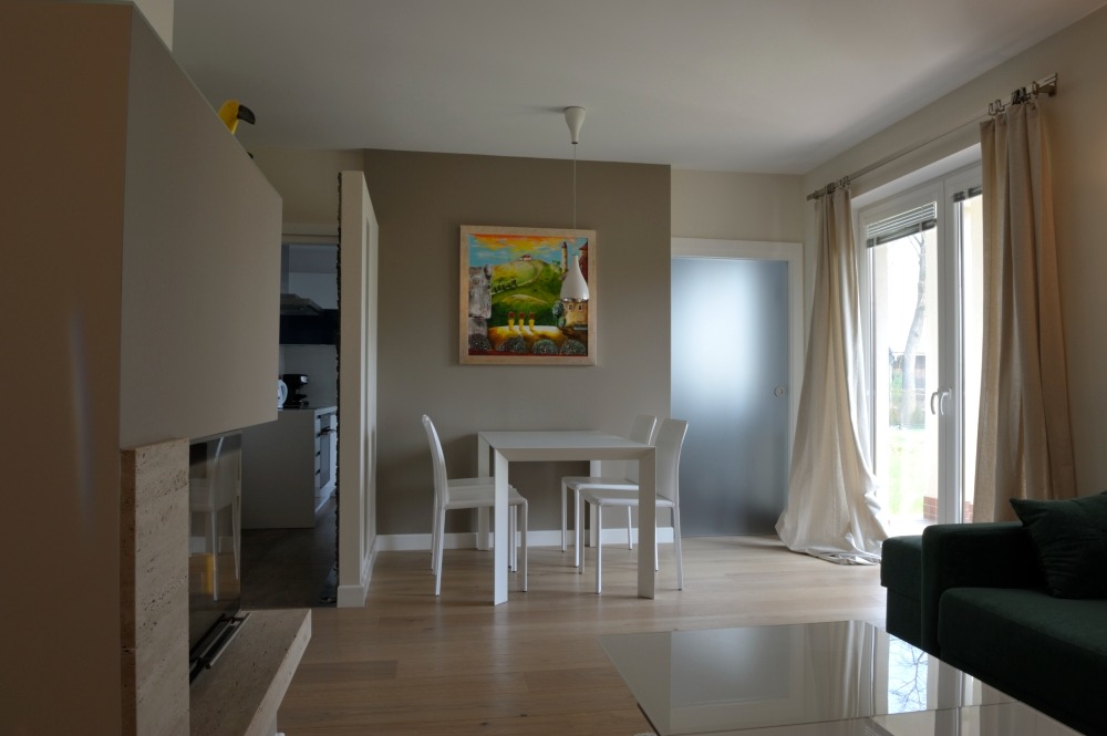 Jasne wnętrze mieszkania z drewnianą podłogą, białym stołem i krzesłami, obrazem na ścianie i widokiem na kuchnię w tle