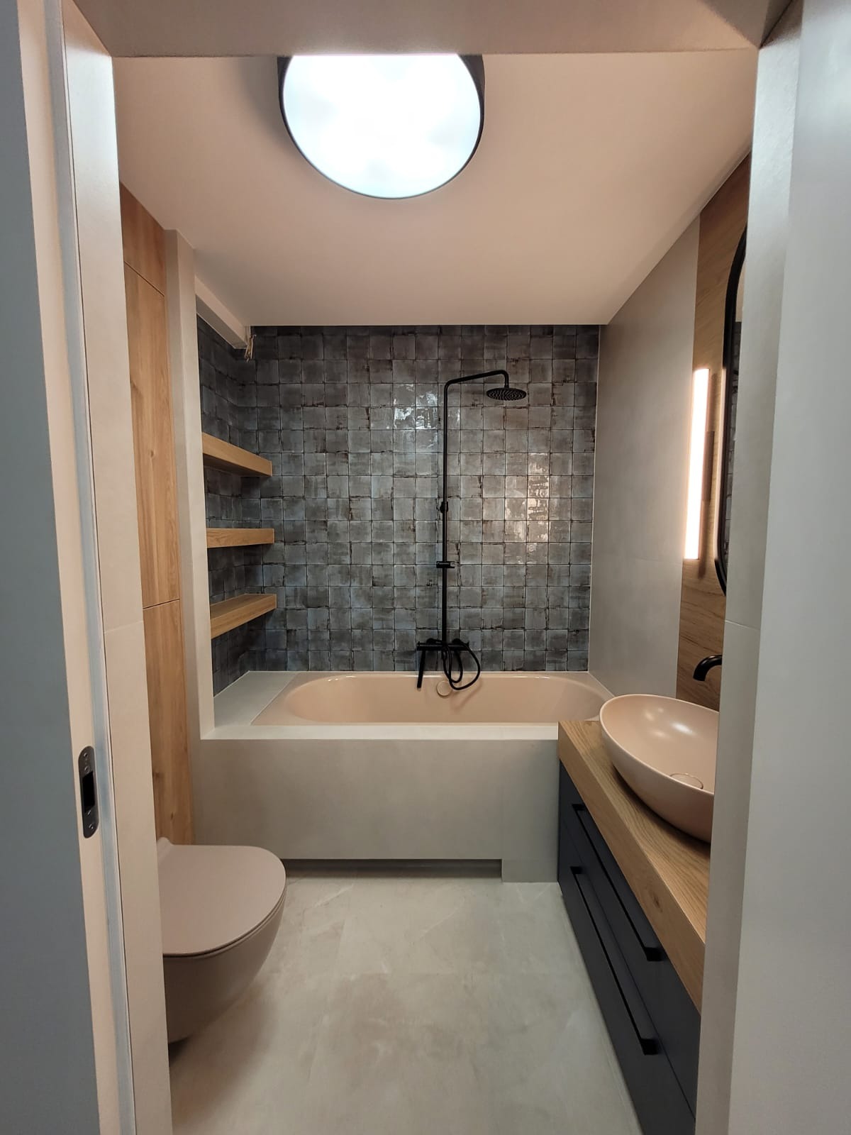 Nowoczesna łazienka z wanną w zabudowie, czarną armaturą i umywalką w odcieniach beżu, oświetlona okrągłym plafonem i pionowym panelem LED.