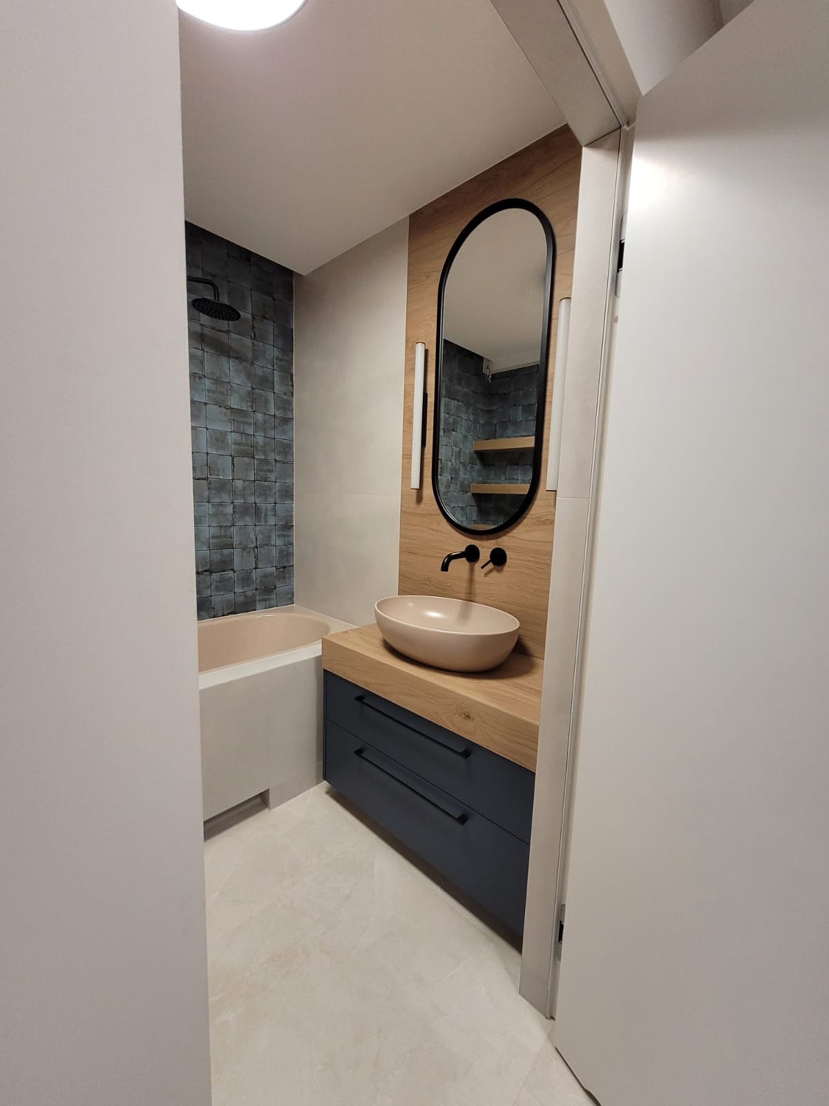 Nowoczesna łazienka z owalnym lustrem, umywalką nablatową w odcieniach beżu i szafką w kolorze granatowym z drewnianym blatem, widoczna kabina prysznicowa z ciemną mozaiką.