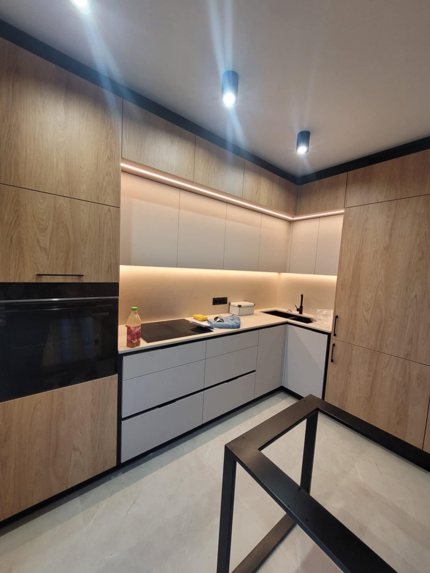 Nowoczesna kuchnia z drewnianymi frontami, szarymi szufladami i oświetleniem LED pod szafkami wiszącymi. Na blacie stoi butelka napoju i gąbka. Widoczny fragment metalowej konstrukcji stołu.