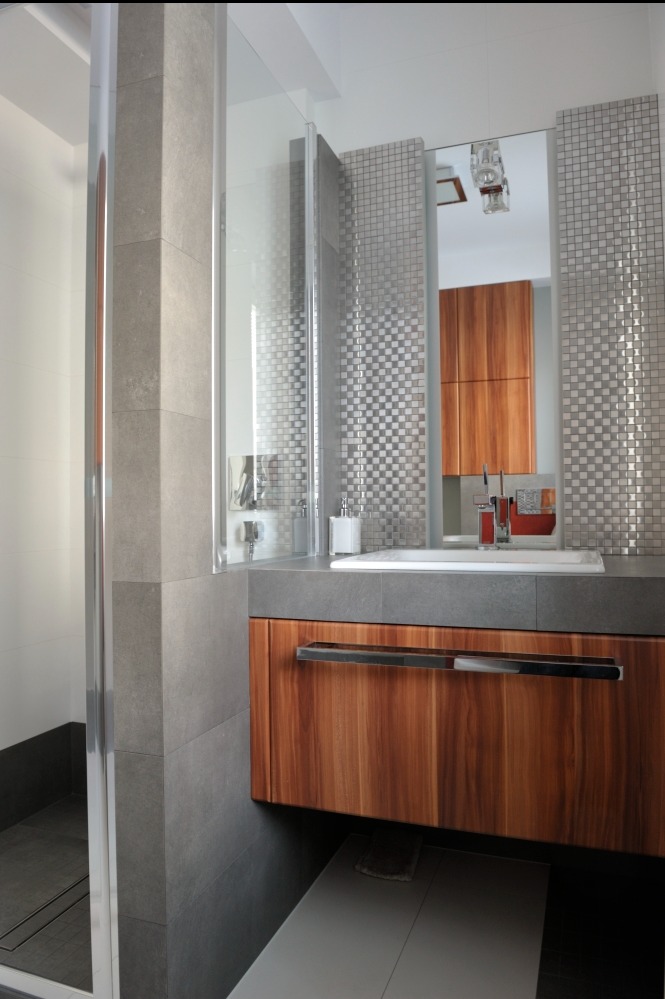 Nowoczesna łazienka z kabiną prysznicową za szkłem, umywalką na szafce z drewnianym frontem i mozaiką na ścianie. Minimalistyczny design, szare płytki i chromowane detale.