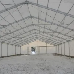 Wnętrze dużej, tymczasowej hali z metalową konstrukcją i białym poszyciem, ziemia pokryta śniegiem, widok na zewnątrz przez otwarte wrota.