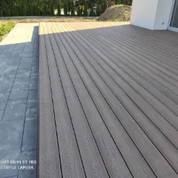 Nowoczesny taras z desek kompozytowych w odcieniach szarości, przylegający do kostki brukowej i budynku z białymi ścianami. W tle widoczne zielone tuje.