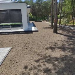 Przygotowany teren pod trawnik w Mniszkowie, z widoczną świeżo nawiezioną ziemią, obrzeżami z kostki brukowej i nowoczesnym domem w tle. W oddali widoczny pracujący sprzęt budowlany.