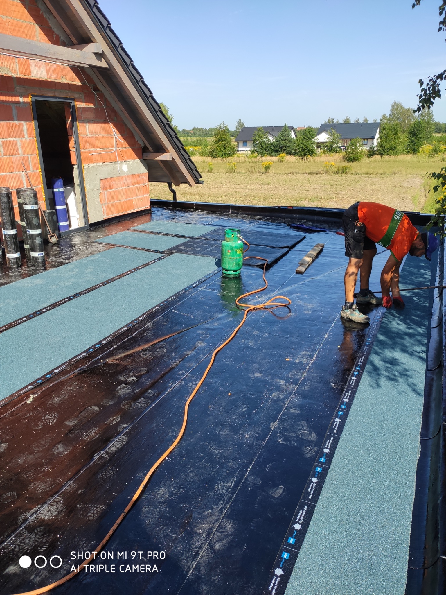 Pracownik w pomarańczowej koszulce i czarnych spodenkach układa papę termozgrzewalną na dachu budynku w trakcie budowy. Widoczna butla gazowa, palnik i rolki papy. W tle krajobraz z domami.
