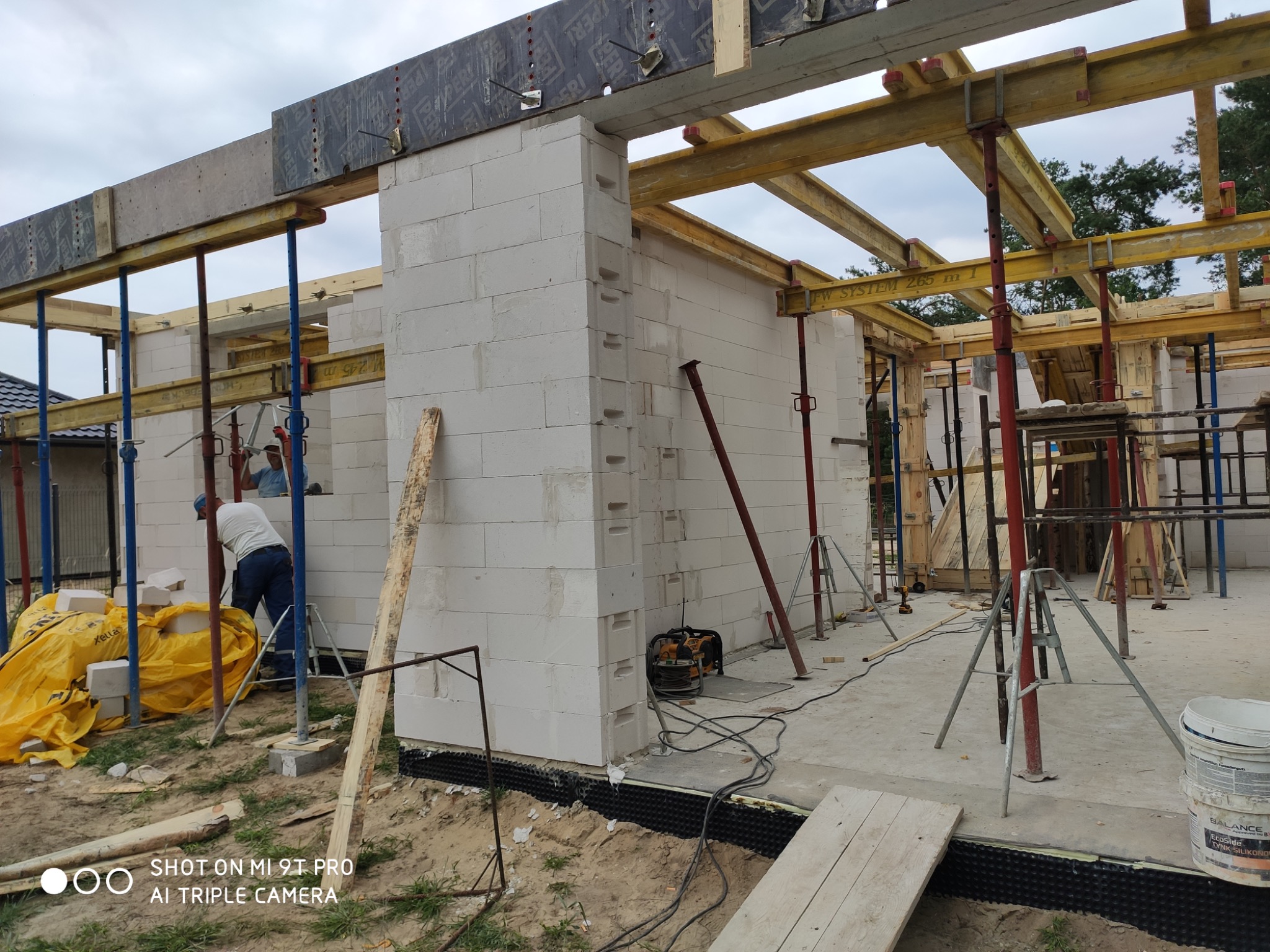 Budowa domu z białych bloczków Ytong, widoczne szalunki, podpory, pracownik w tle oraz narzędzia na placu budowy.