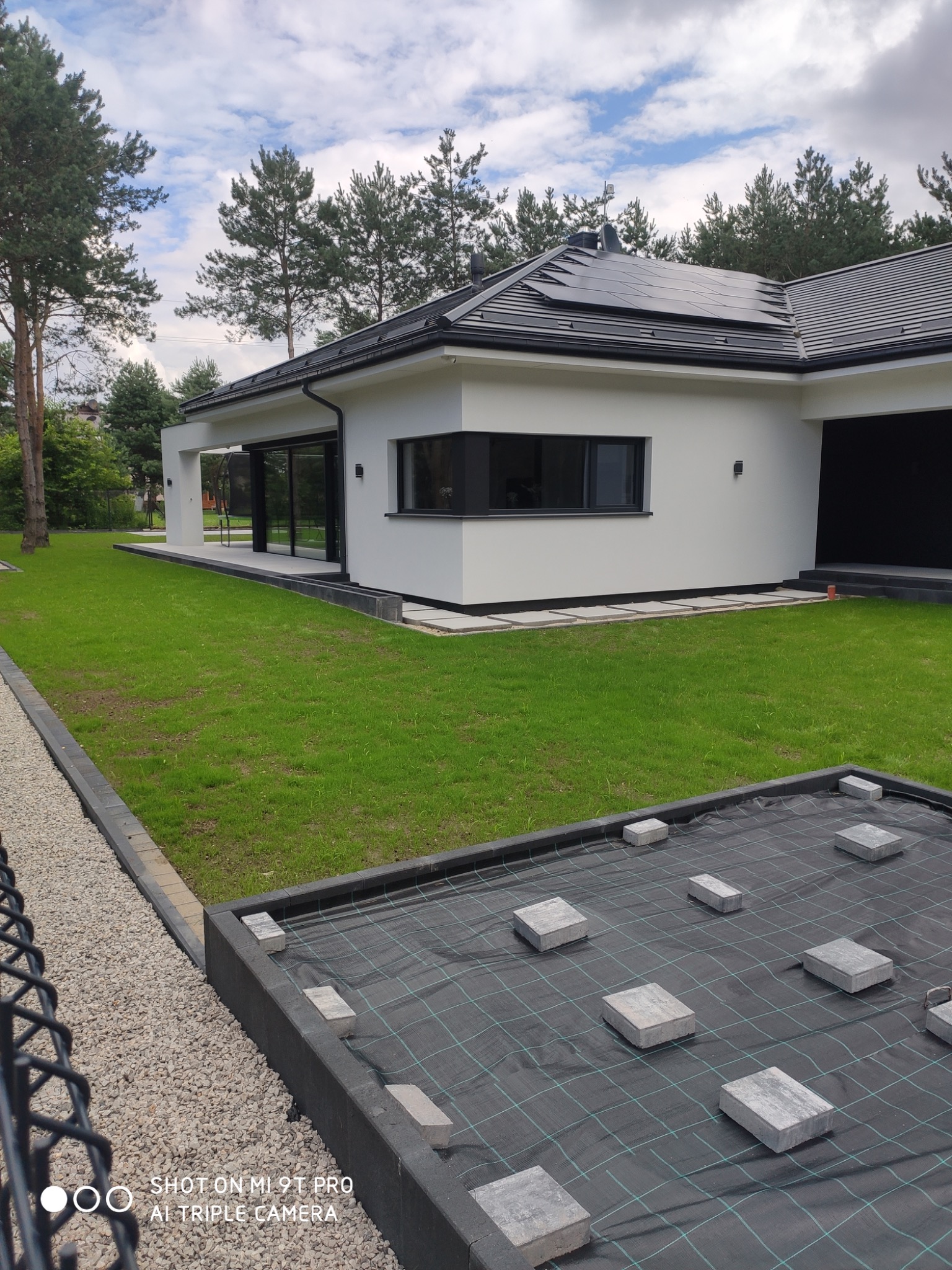 Nowoczesny dom jednorodzinny z panelami słonecznymi na dachu, widok na trawnik oraz przygotowany teren pod kostkę brukową z rozłożoną agrowłókniną i ułożonymi kostkami w równych odstępach.