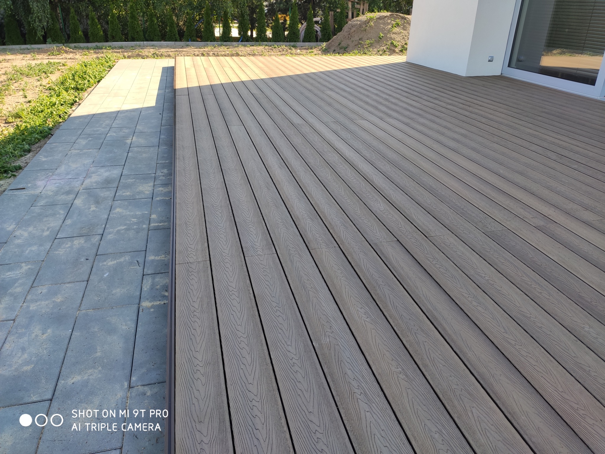 Nowoczesny taras z desek kompozytowych w odcieniach szarości, przylegający do kostki brukowej i budynku z białymi ścianami. W tle widoczne zielone tuje.