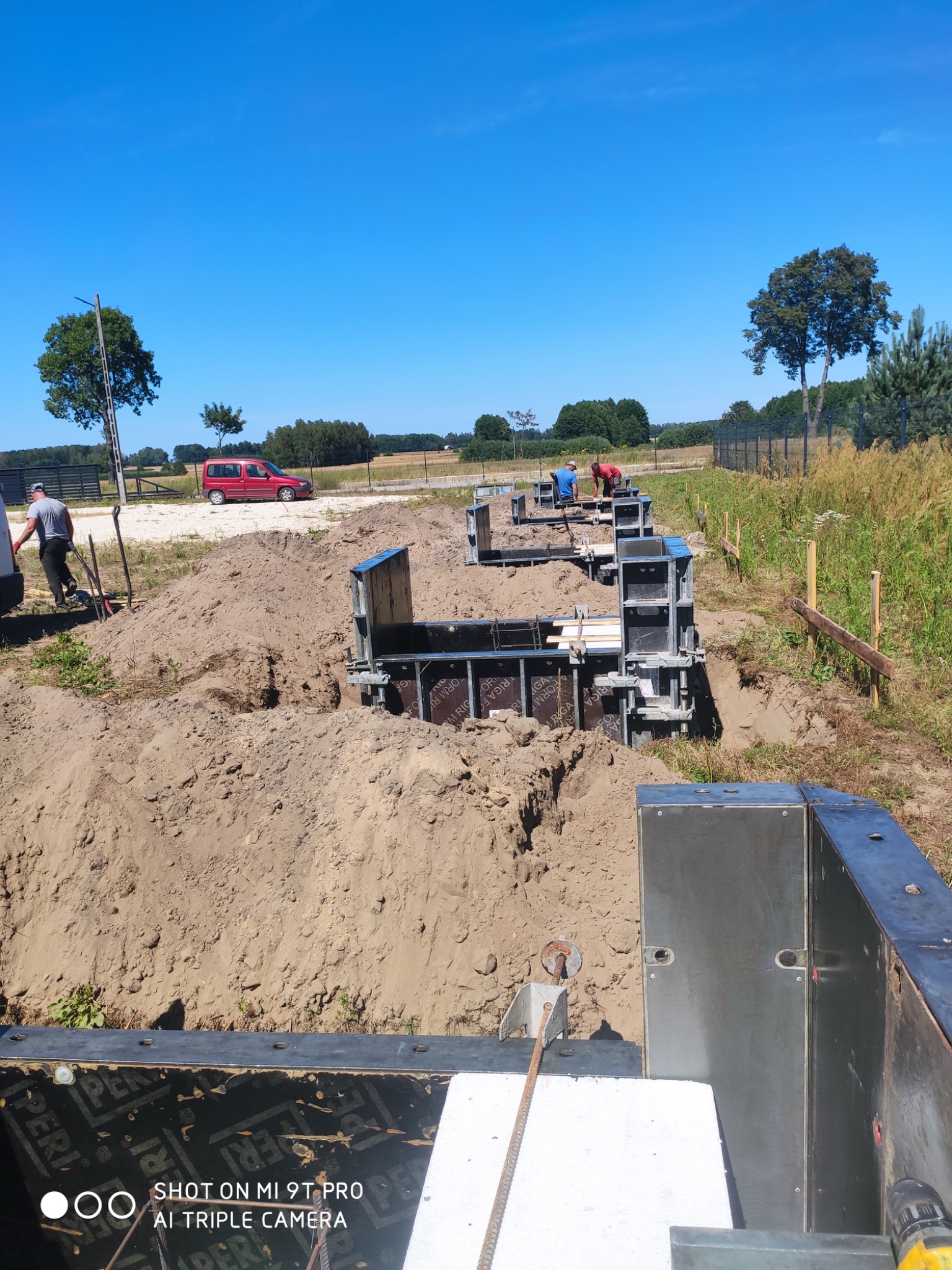 Fundamenty szalunkowe w wykopie, widoczni pracownicy budowlani, krajobraz polny w tle, błękitne niebo, furgonetka w oddali.