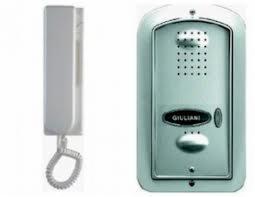 Zestaw domofonowy: słuchawka w kolorze białym i panel zewnętrzny z obudową w kolorze srebrnym, z widocznym przyciskiem dzwonka i mikrofonem.