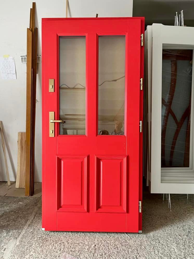 Czerwone drzwi z dwoma szklanymi panelami, zamontowane okucia w kolorze złotym, widoczne odbicie osoby w szkle, obok białe drzwi w folii ochronnej, przygotowane do montażu.
