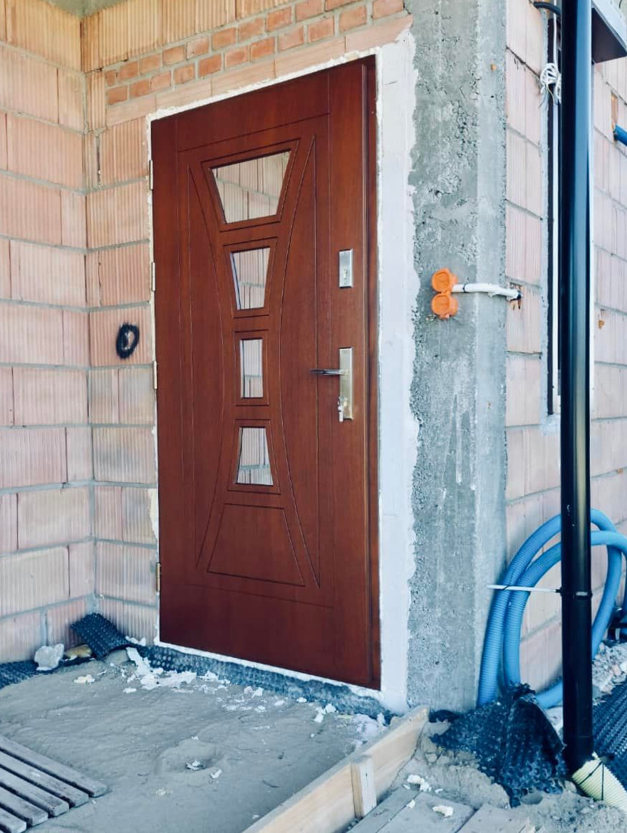 Brązowe drzwi wejściowe z szybami w kształcie trapezu, osadzone w surowym murze z cegły i betonu, widoczne elementy instalacji elektrycznej i niebieski wąż, stan surowy.