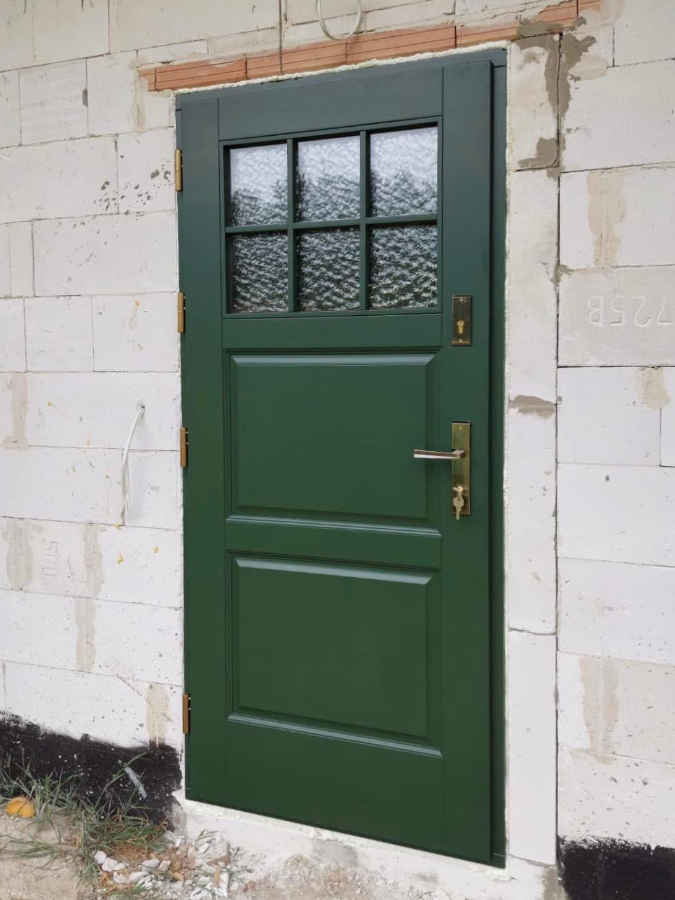 Zielone, częściowo przeszklone drzwi wejściowe, zamontowane w surowej ścianie z białych bloczków betonowych, widoczne niedokończone obróbki murarskie.