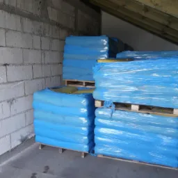 Stosy worków z pelletem na drewnianych paletach, owinięte w niebieską folię, ustawione w magazynie z surowymi ścianami z betonowych bloków.