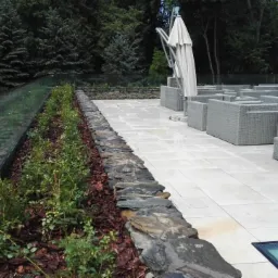 Nowoczesny taras z jasną kostką, szarymi meblami rattanowymi, szklaną balustradą i rabatą z krzewami ozdobnymi, wykończoną korą.