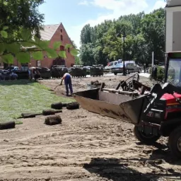 Układanie trawy z rolki: Pracownik wyrównuje teren, w tle budynki i zwinięte rolki trawy, na pierwszym planie traktor z łyżką.