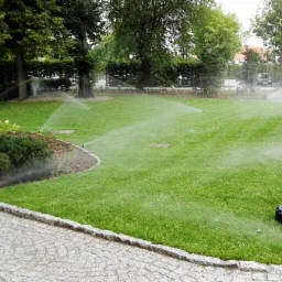 Automatyczny system nawadniania trawnika w ogrodzie, zraszacz trawnikowy w trakcie pracy, widoczna dysza i strumień wody, równomiernie nawodniona trawa, dekoracyjna lampa ogrodowa w tle.