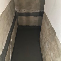 JUSTYNA JABŁOŃSKA BUDOWNICTWO - Świeżo zaizolowany fundament w Łodzi, z widocznymi warstwami hydroizolacji na ścianach i podłodze. Szare ściany i podłoże, widoczne ślady po uszczelnieniach.