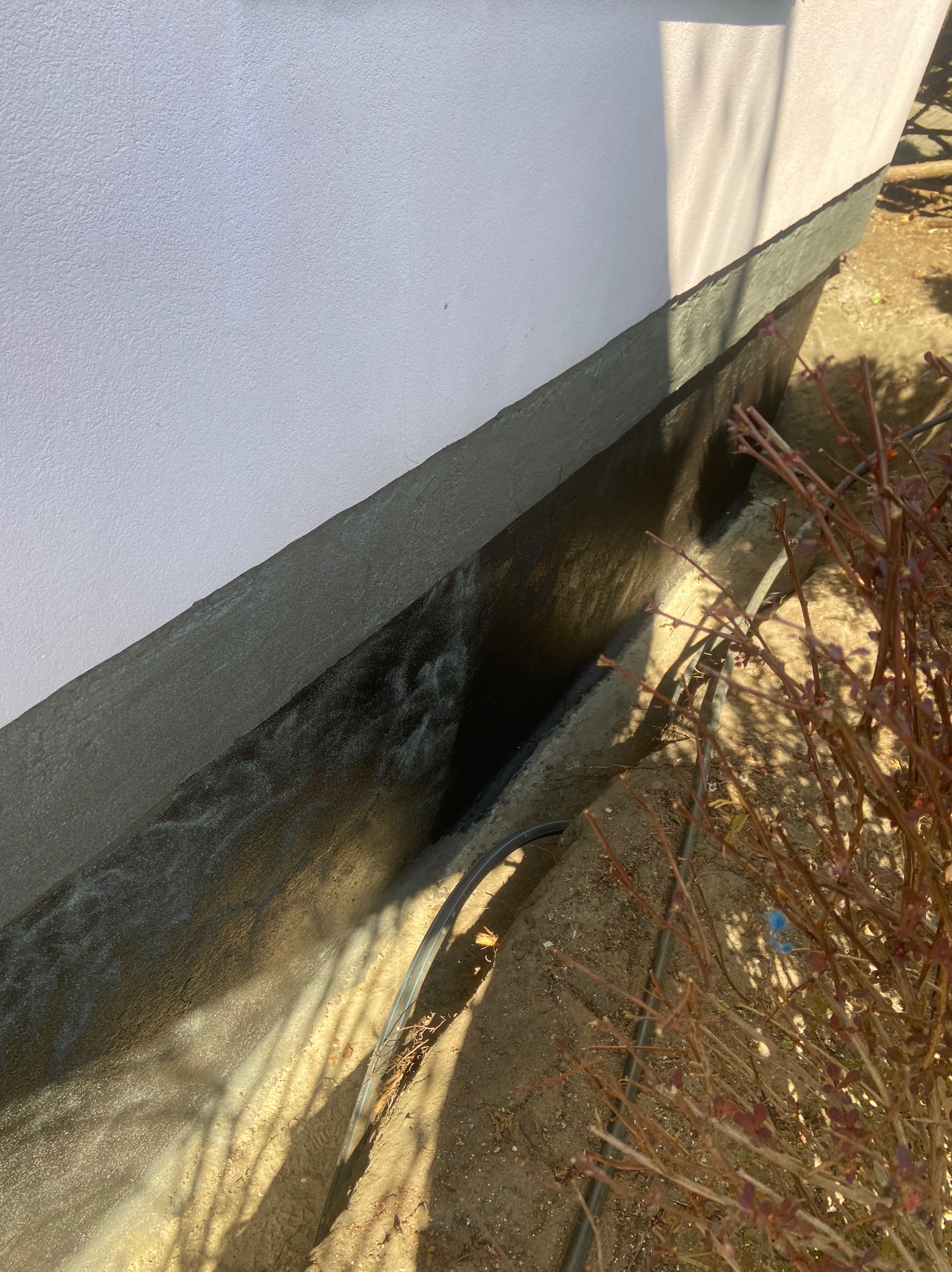 Wykończona izolacja fundamentu budynku z widoczną czarną powłoką hydroizolacyjną i szarym pasem ochronnym, na tle białej ściany i ziemi.