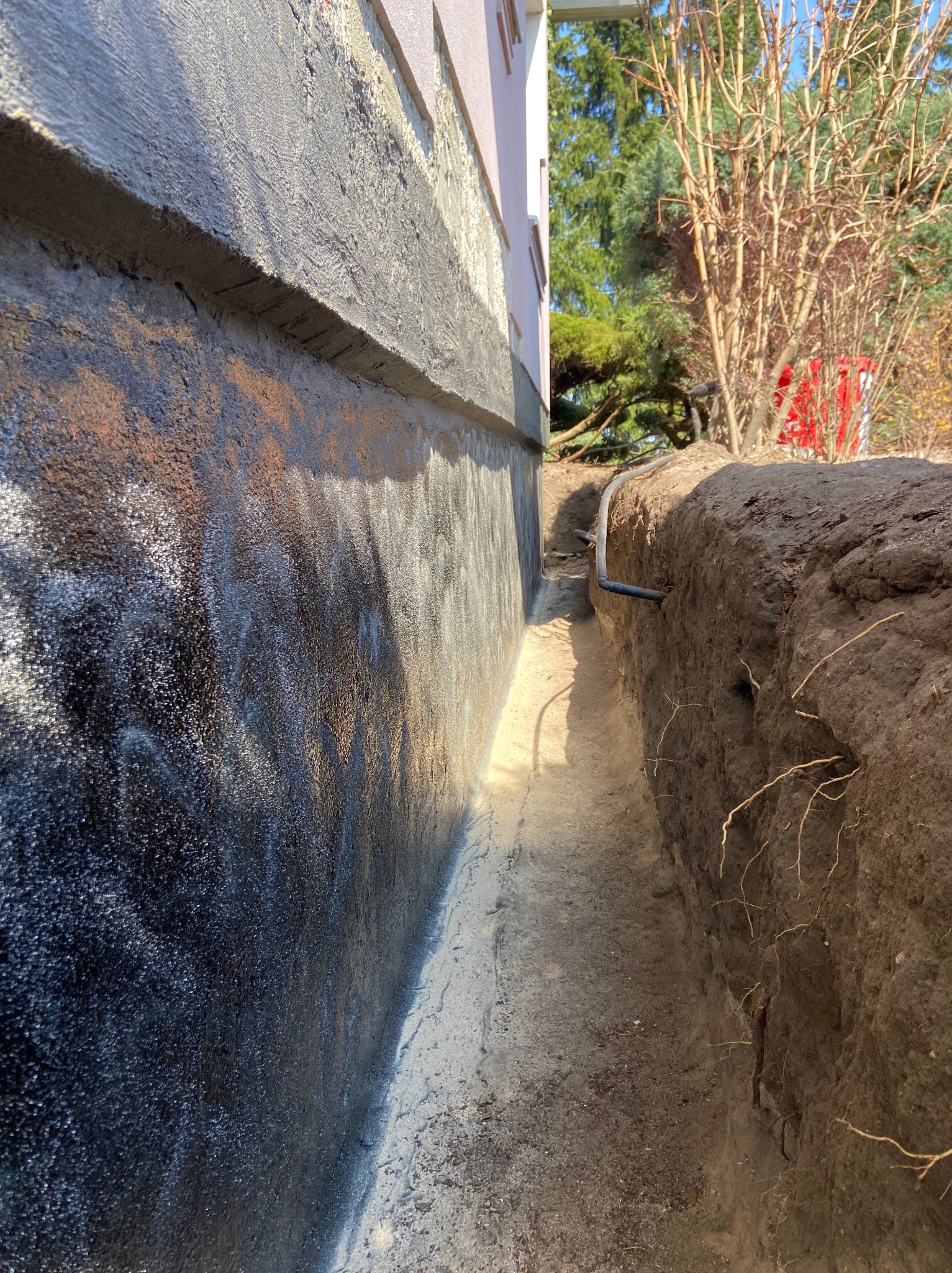 Fundament budynku po izolacji, z wykopem i widoczną rurą drenażową. Czarna, błyszcząca powłoka izolacyjna na ścianie fundamentu.