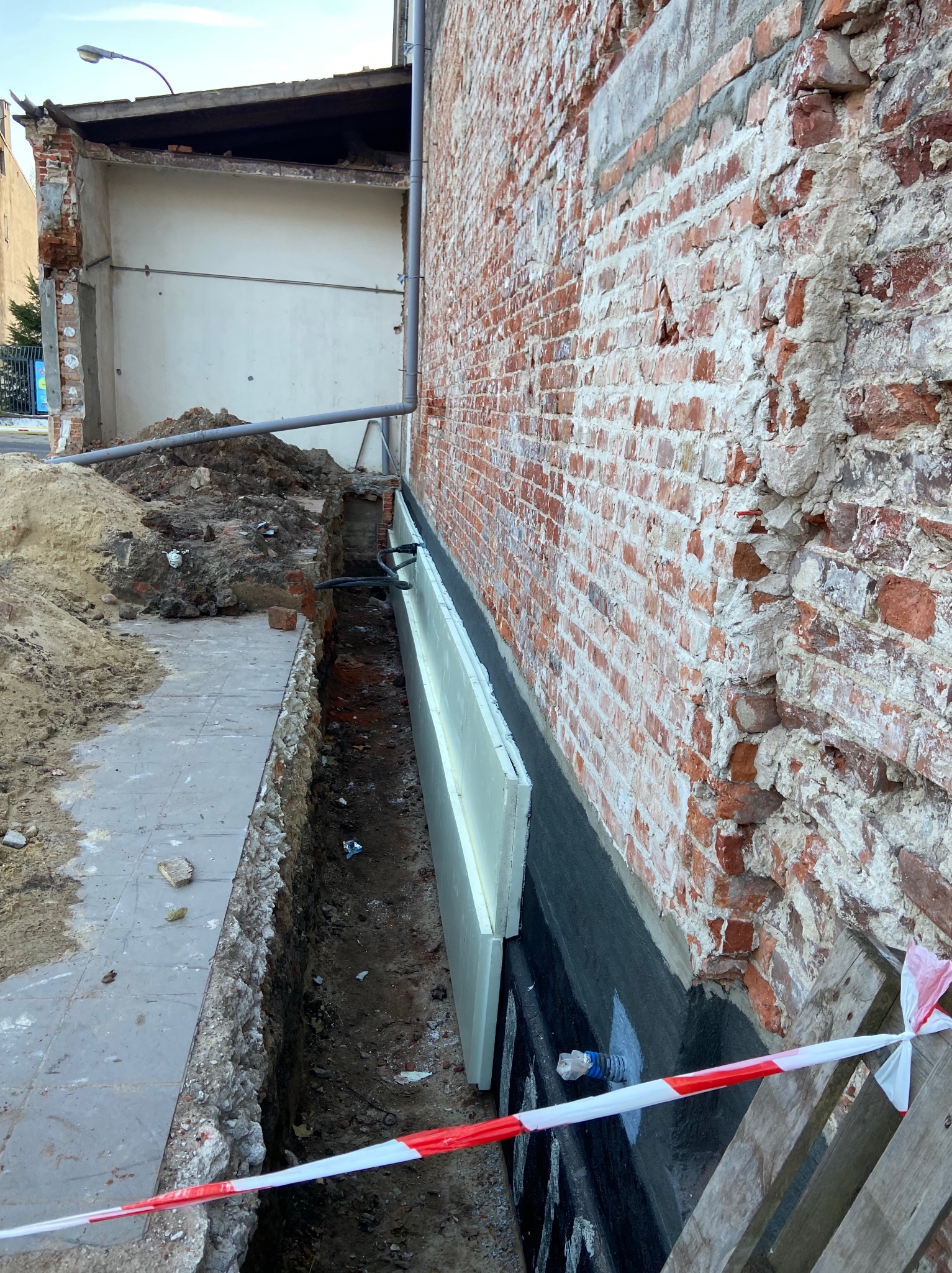 Odkopane fundamenty ceglanego budynku z widoczną izolacją termiczną (styropian) i hydroizolacją (czarna folia). Wykop zabezpieczony taśmą ostrzegawczą.