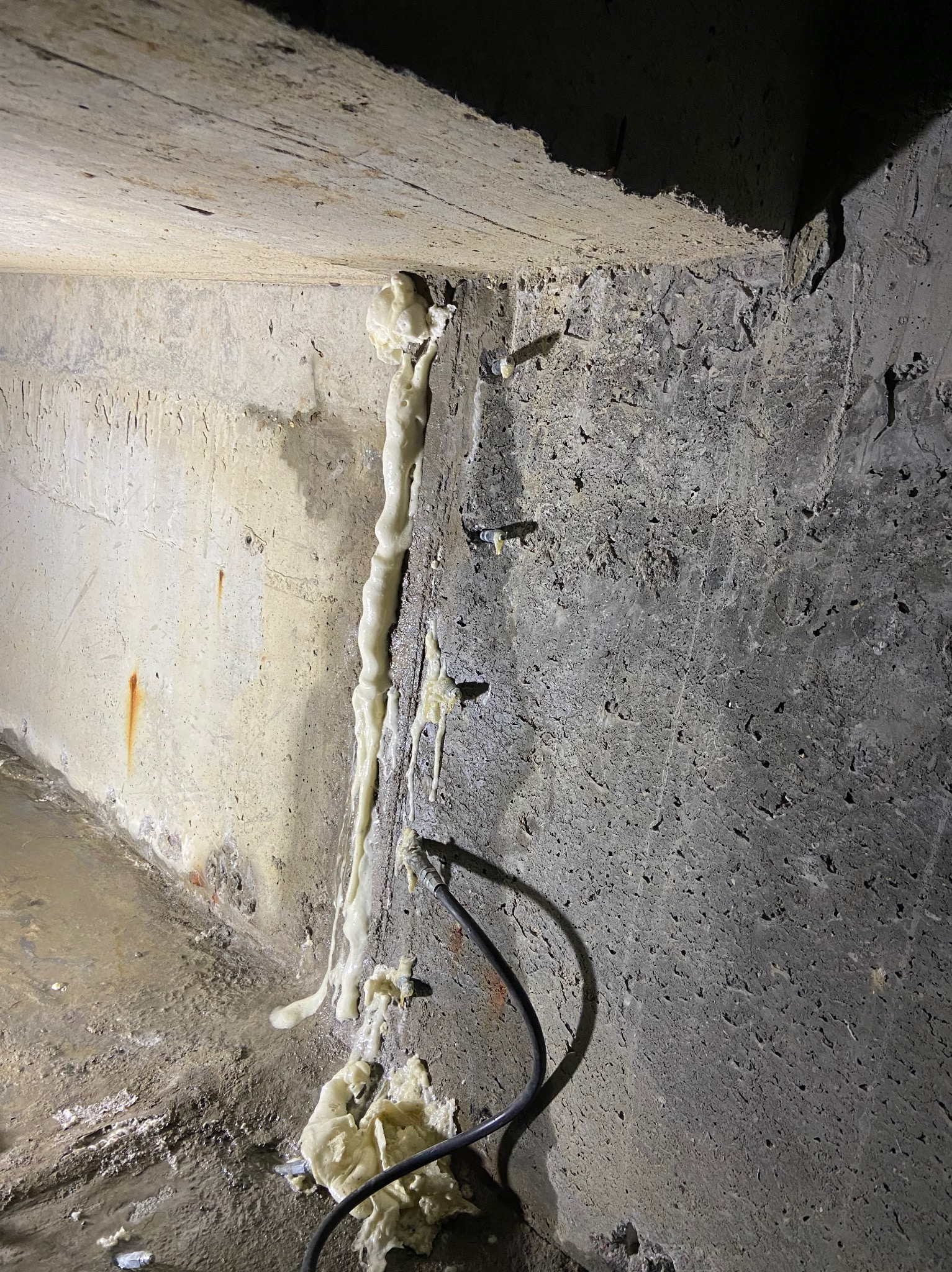 Szczegółowe ujęcie izolacji pianką montażową w szczelinie betonowej ściany fundamentowej z widocznymi kotwami. Przewód w dolnej części kadru.