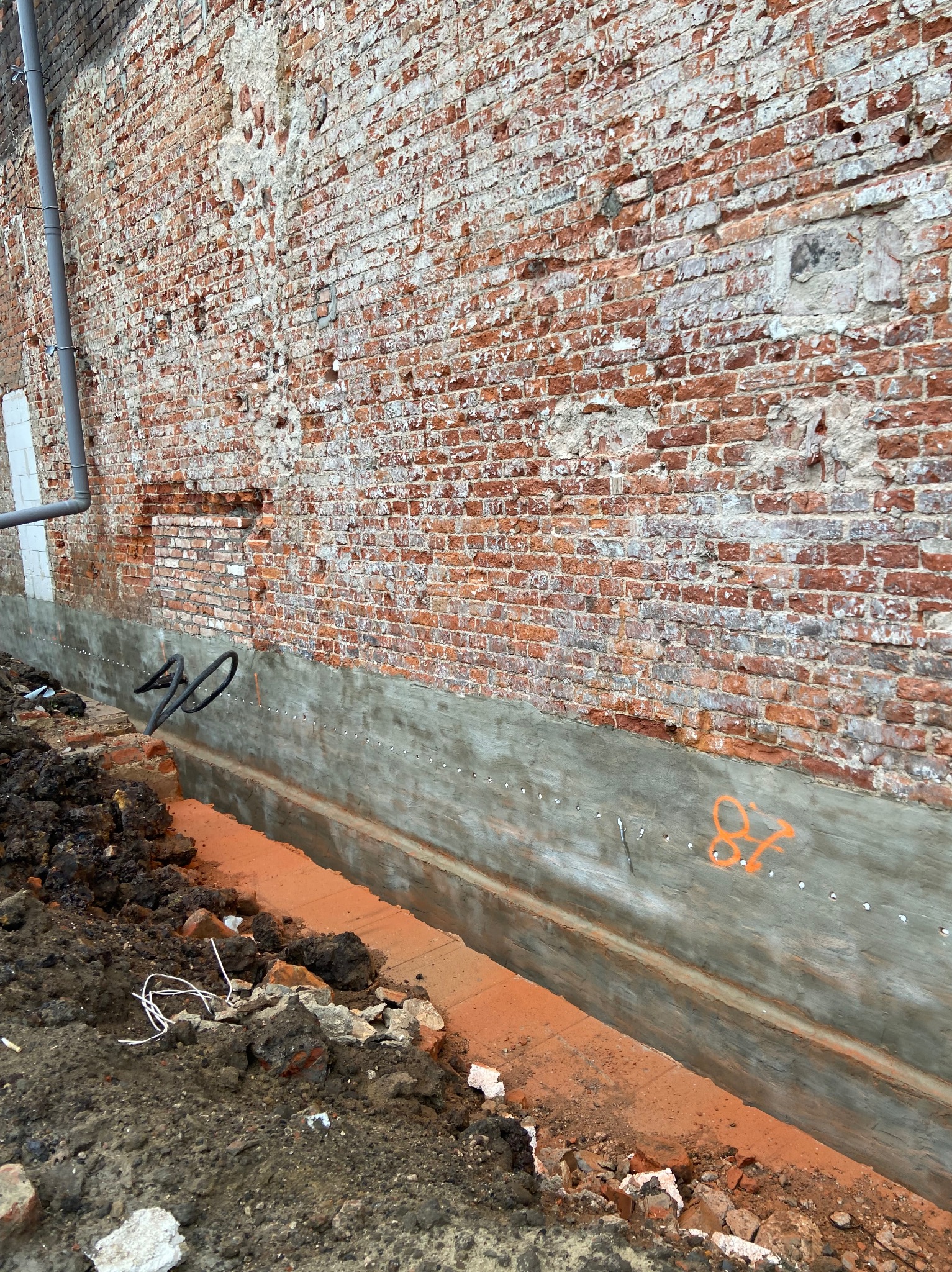 Odkopany fundament ceglanego budynku z widoczną warstwą betonu i pomarańczową substancją izolacyjną w wykopie. Rura spustowa wzdłuż ściany.