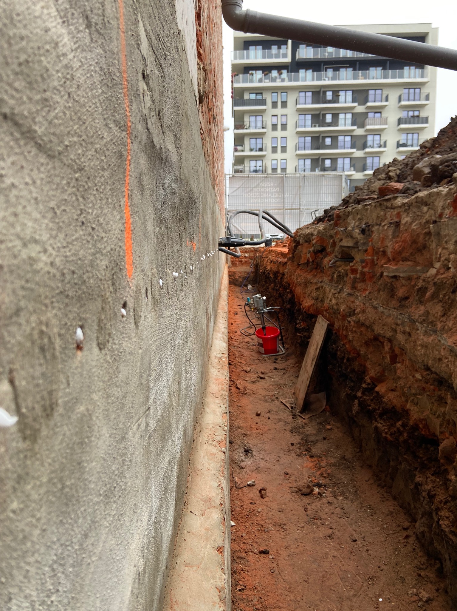 Wykop wzdłuż ściany budynku z widocznymi punktami iniekcji i sprzętem do osuszania. W tle budynek mieszkalny.