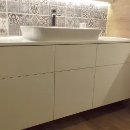 Biała szafka łazienkowa z umywalką nablatową i baterią, tło z płytek patchworkowych i drewnopodobnych, widoczne gniazdko elektryczne.