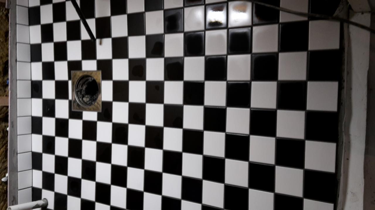 Świeżo ułożona, czarno-biała szachownica z płytek ceramicznych na podłodze prysznica, z metalowym odpływem w rogu, widoczne niedociągnięcia na krawędziach i pozostałości zaprawy.