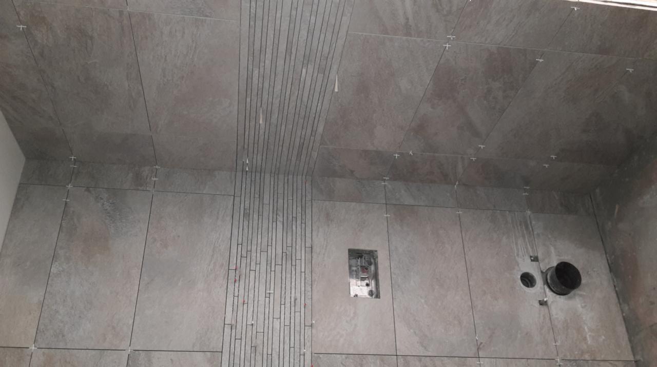 Układanie płytek w łazience: szare płytki imitujące kamień na ścianach i suficie, z dekoracyjnym pasem pionowych, wąskich płytek oraz widocznymi krzyżykami dystansowymi i otworami na instalacje.