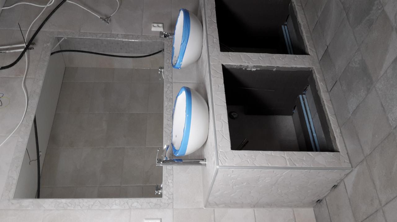 Dwie białe umywalki z niebieską taśmą ochronną, zamontowane w łazience w trakcie wykończenia, widoczne otwarte przestrzenie szafek pod umywalkami, szare płytki na ścianach i podłodze.