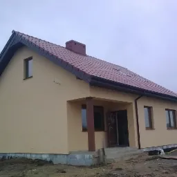 Gotowy, otynkowany dom jednorodzinny z czerwonym dachem, widoczny od strony frontowej z podcieniem wejściowym i schodami, położony na działce w pochmurny dzień.