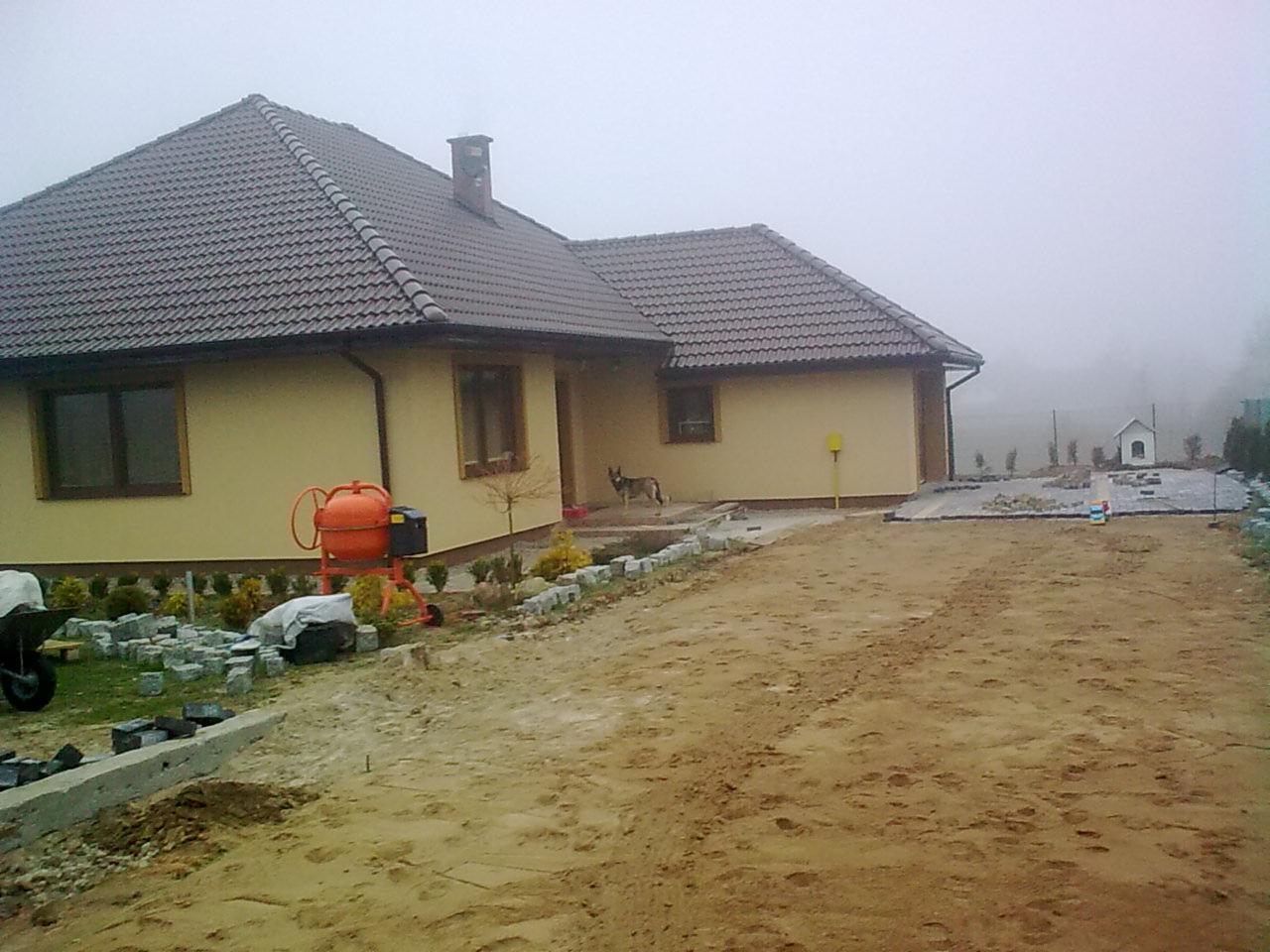 Parterowy dom z brązowym dachem w trakcie budowy pod klucz, widoczna pomarańczowa betoniarka, ułożone kostki brukowe i pies w wejściu, mglista pogoda.